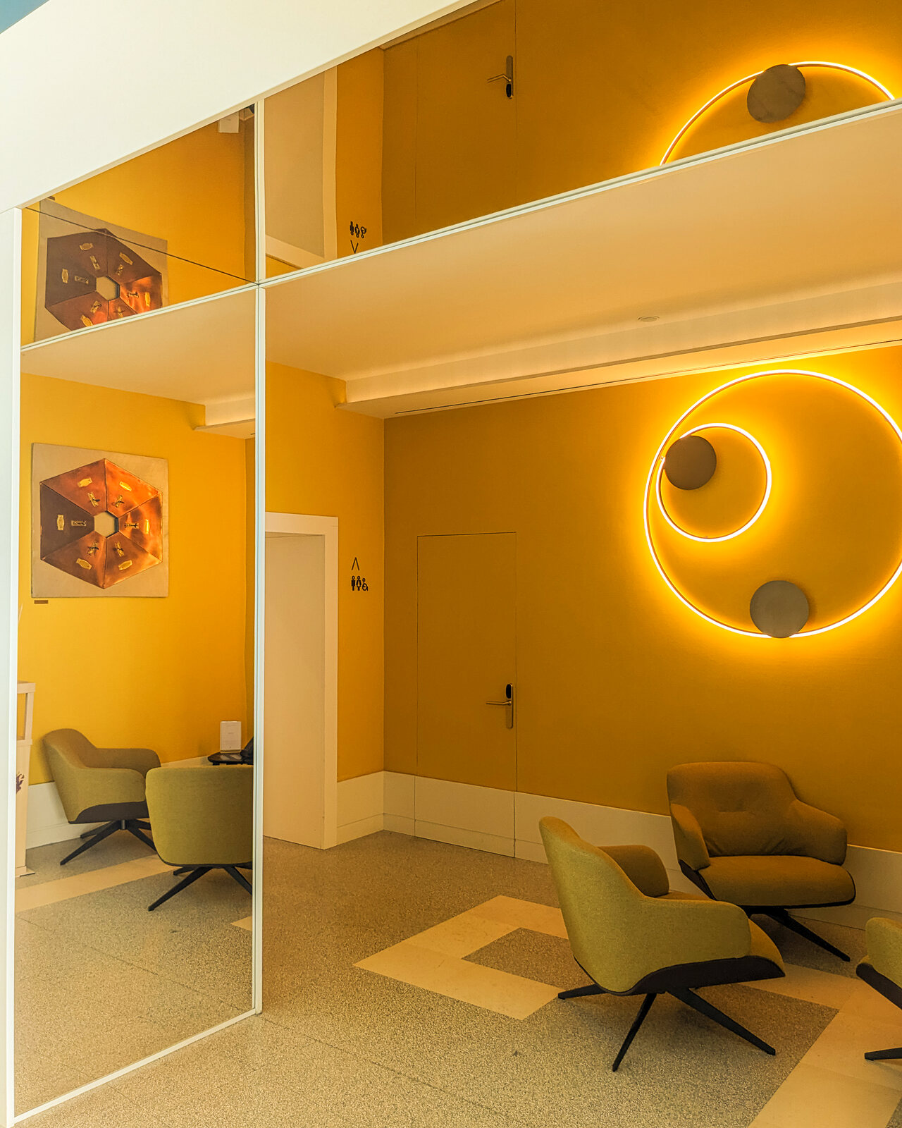 Mid-century lounge with mustard-yellow walls, a circular neon light, and green armchairs reflected in a mirrored wall.