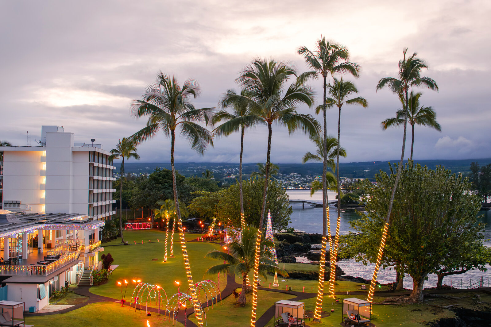 Oceanfront resort grounds at dusk with palm trees wrapped in string lights and the sea beyond.