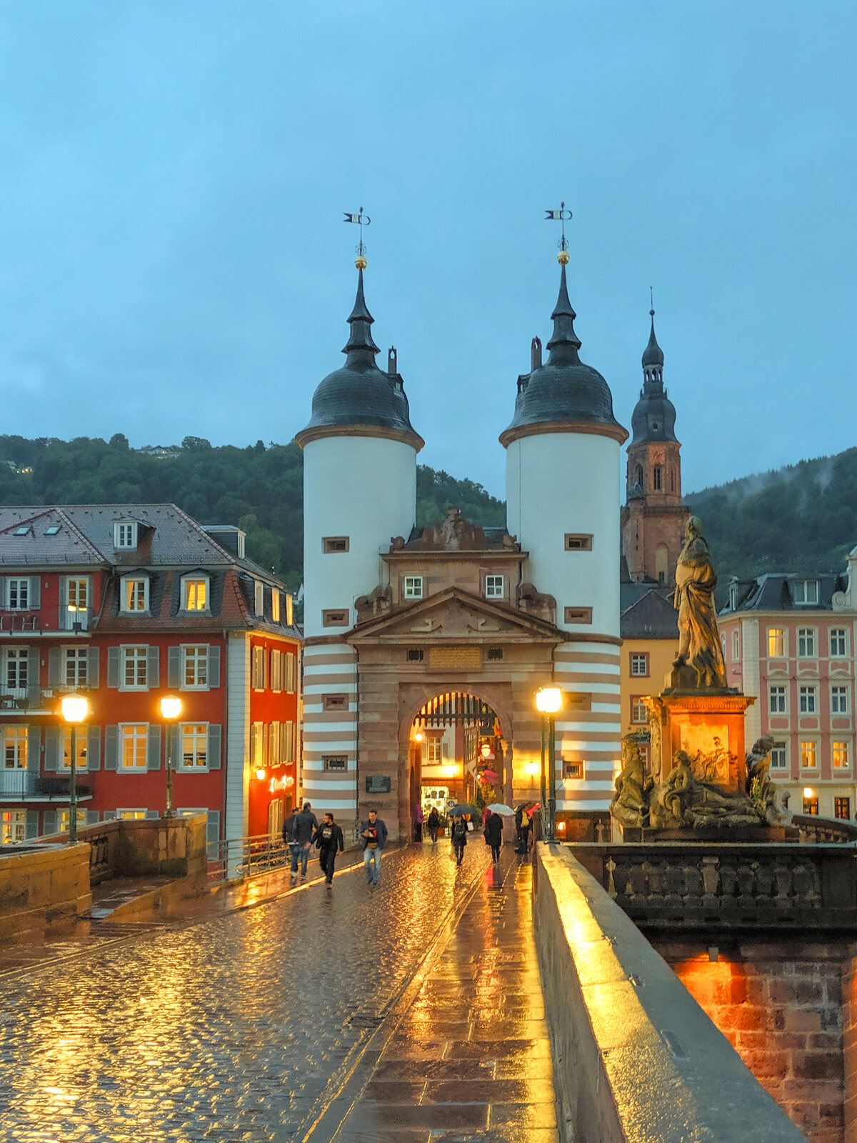 Heidelberg's Old Bridge gate with twin towers at blue hour, reflected in rain-slicked cobblestones.